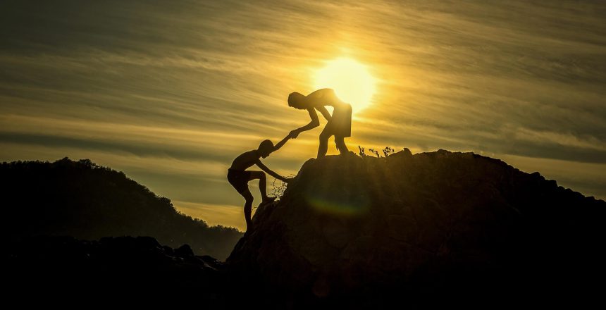 Henkilö auttaa toista kiipeämään kukkulalle. Taustalla näkyy auringonlasku.